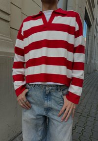 Sarkani un baltu svītrainu garroku polo kreklu ar V veida izgriezumu, saskaņotu ar gaiši ziliem džinsiem no džinsa, demonstrējot brīvu piegriezumu un ikdienišķu stilu.