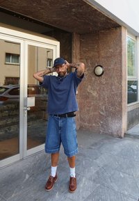 Mand med tatoveringer iført blå kasket, mørkeblå skjorte, denimshorts, hvide sokker og brune sko står med hænderne bag hovedet nær glasdøren.