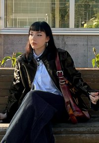 Giacca di pelle nera, camicia bianca e gonna in denim scuro. Borsa marrone con hardware metallico. Capelli neri lunghi e lisci, seduta su una panchina.