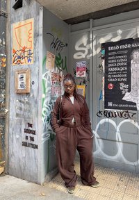 A young person with braided hair and wearing a brown outfit is leaning against a wall covered in graffiti, surrounded by event posters in an urban environment.
