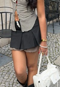Schwarzer plissierter Rock über einem karierten Minirock, kombiniert mit einem karierten Crop-Top. Die Accessoires umfassen eine weiße Handtasche und perlenartige Perlen am Bund.