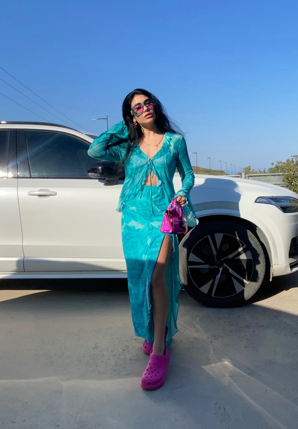 Turquoise two-piece outfit comprising a cropped sheer top and a long skirt with a thigh-high slit, paired with pink clogs and a small shiny handbag.