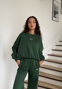 Femme aux cheveux bouclés portant un sweat-shirt Nike vert foncé et un pantalon, debout à l'intérieur près d'un escalier en bois et de photos encadrées en noir et blanc.