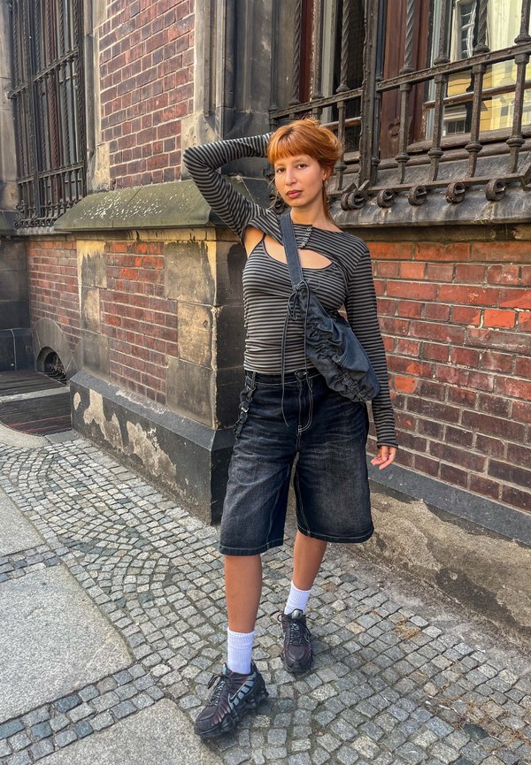 Ung kvinde med kort rødt hår iført en stribet top med udskæringer, sorte shorts, sneakers og en crossbody taske står på en brostensbelagt gade ved en murstensbygning.