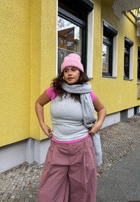 Haut gris sans manches porté sur un t-shirt rose, associé à un pantalon rose ample. Un foulard gris et un bonnet rayé rose complètent la tenue. Fond mural jaune.