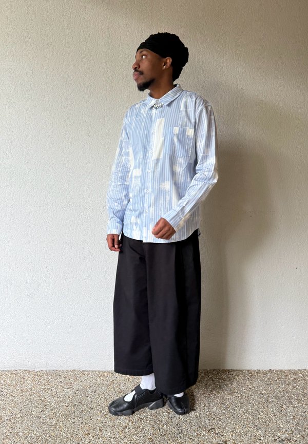 Homme portant une chemise à manches longues bleu clair à motif, un pantalon noir à jambes larges, des chaussettes blanches, des chaussures noires et un foulard noir sur la tête, debout contre un mur.