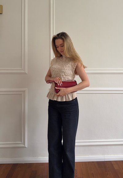 Mujer con top sin mangas beige texturizado y pantalones negros, sosteniendo un pequeño bolso rojo, de pie frente a una pared blanca con molduras decorativas.