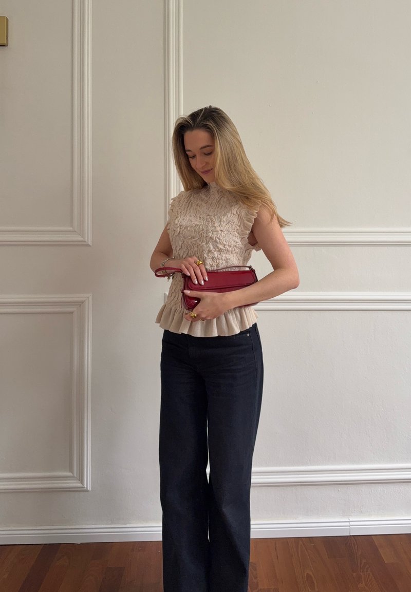 Femme en haut sans manches beige texturé et pantalon noir, tenant une petite pochette rouge, debout contre un mur blanc avec moulures décoratives.