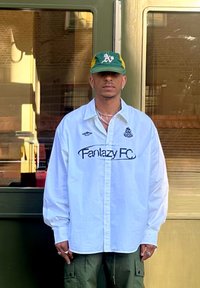 Fehér, hosszú ujjú ing, rajta a "Fantazy FC" felirattal, zöld és sárga baseball sapkával, valamint olíva zöld cargo nadrággal párosítva. Ezüst ékszerek kiegészítik az outfitet.