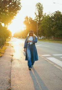 Casaco de denim e jeans de perna larga em azul, camisa branca e boné castanho. A carregar uma bolsa branca, a caminhar numa rua iluminada pelo sol, ladeada de árvores.