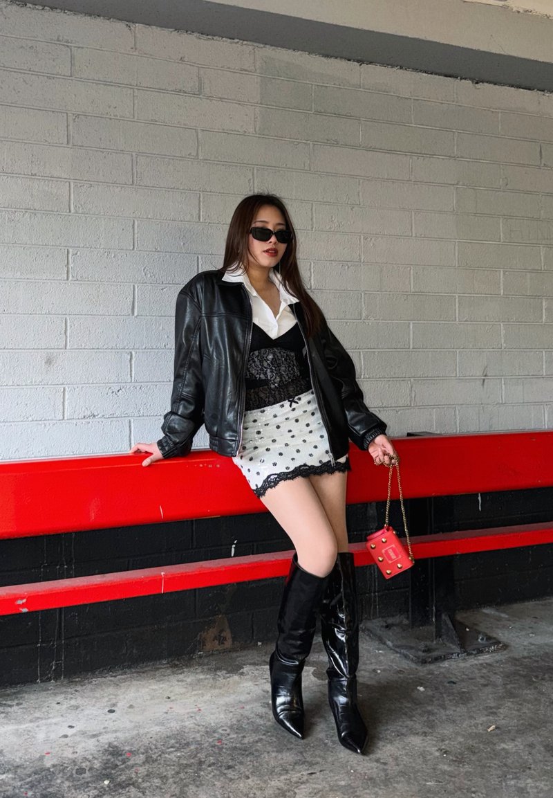 Junge Frau in schwarzer Lederjacke, weisser Bluse, gemustertem Rock, schwarzen kniehohen Stiefeln und Sonnenbrille lehnt drinnen an einem roten Geländer.