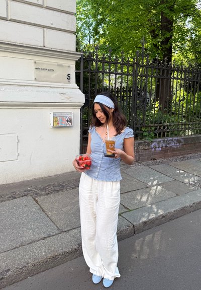 Una mujer con una blusa de cuadros azul y pantalones de lino blancos sostiene un recipiente de fresas y una bebida, de pie junto a un edificio con árboles verdes.