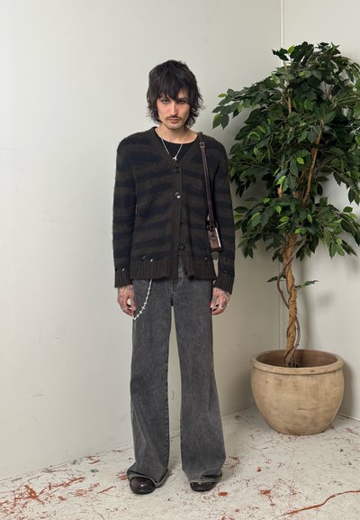 Joven con cabello despeinado que lleva un cárdigan negro con rayas, pantalones vaqueros grises anchos, bolso cruzado y accesorio de cadena, de pie junto a una maceta grande con planta.