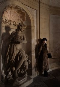 Personne portant un manteau à imprimé léopard se tient à côté d'une statue classique en pierre d'une femme semi-nue tenant un pichet dans une niche architecturale vieillie.