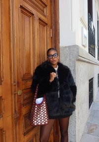 Μαύρο φουσκωτό παλτό με ψηλό γιακά, συνδυασμένο με μια μπορντό τσάντα tote με ασημένιες κουκίδες. Ξύλινη πόρτα χρησιμεύει ως φόντο.