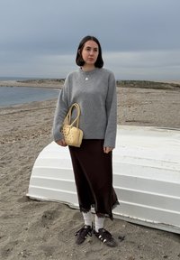 Γυναίκα που στέκεται σε παραλία κοντά σε αναποδογυρισμένο λευκό σκάφος, φοράει γκρι πουλόβερ, σκούρα φούστα, αθλητικά παπούτσια και κρατάει μια μπεζ τσάντα.