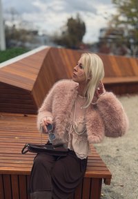 Lyserød faux fur jakke med brede ærmer, lagdelt over en beige blonde top. Mørkebrune bukser, holder solbriller og en sort håndtaske.