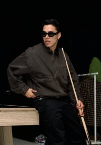 Braune Denimjacke mit Reißverschluss, zwei Fronttaschen, schwarze Cargo-Hosen, hält einen Queue in der Hand, trägt dunkle Sonnenbrille. Schwarzer Hintergrund.