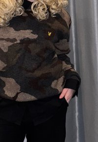 Pull en maille à motif camouflage dans des tons de brun, gris et noir, avec un petit logo jaune et des poignets noirs. Porté sur une chemise noire.