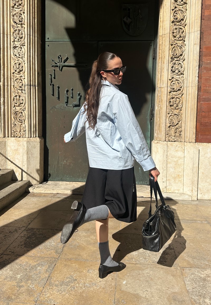 Donna con capelli lunghi, occhiali da sole e camicia oversize solleva una gamba, tenendo una borsa nera vicino a un ornato ingresso in pietra al sole.