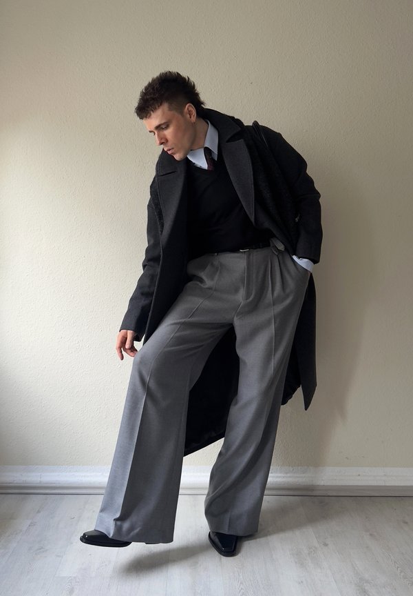 Man die een zwarte jas, zwart trui, wit overhemd en brede grijze broek draagt, voorover leunend met één hand in de zak tegen een effen muur.