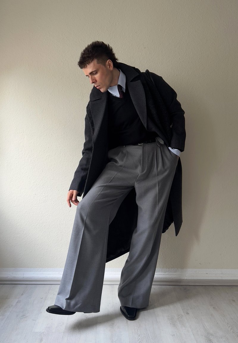 Mann in schwarzem Mantel, schwarzem Pullover, weißem Hemd und weiten grauen Hosen, der sich mit einer Hand in der Tasche gegen eine schlichte Wand lehnt.