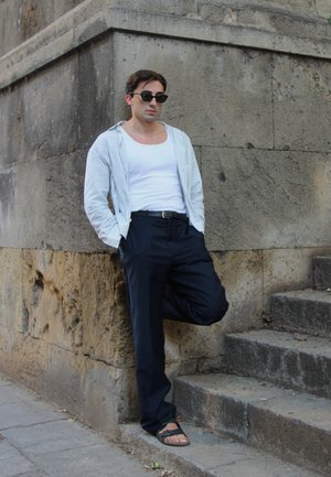 Modelo vistiendo una camiseta sin mangas blanca, una camisa abierta a rayas, pantalones oscuros y chanclas, apoyado contra una pared de piedra cerca de unas escaleras.