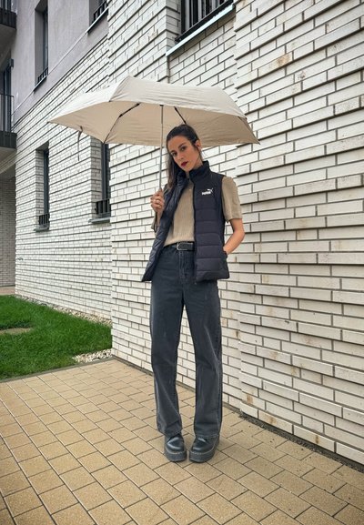 Chaleco acolchado negro sobre una blusa beige de canalé, combinado con pantalones oscuros de pierna ancha y botas negras voluminosas. Sosteniendo un paraguas de color claro.