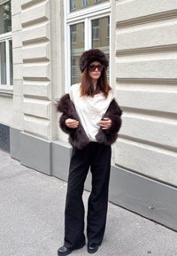 Model poartă un top alb cu decolteu larg și o geacă de blană închisă la culoare. Pantaloni negri cu croială largă și cizme închise la culoare completează ținuta.