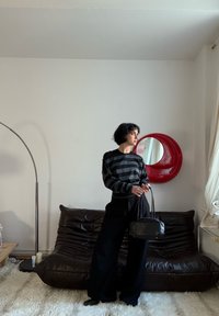 Pull rayé noir et gris, pantalon large noir, tenant un petit sac à main noir. Canapé en cuir marron et miroir circulaire rouge en arrière-plan.