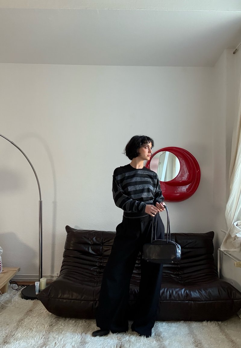 Pull à rayures noires et grises, pantalon large noir, tenant un petit sac à main noir. Canapé en cuir marron et miroir circulaire rouge en arrière-plan.