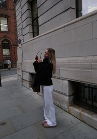 Persoon in een zwarte jas houdt een zonnebril vast voor een licht stenen gebouw. Ze dragen een witte broek en lichtgekleurde instapschoenen.