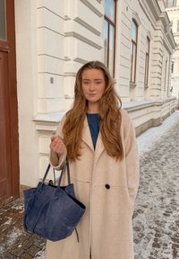 Femme aux longs cheveux bruns portant un manteau beige et tenant un sac à main bleu marine dans une rue enneigée avec des façades de bâtiments historiques en arrière-plan.