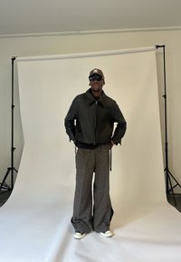 Bărbat îmbrăcat cu o jachetă de piele neagră, pantaloni gri largi, sneakers, ochelari de soare și o șapcă de baseball, stând în fața unui fundal alb.