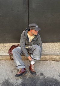 Persona con felpa grigia, jeans e stivaletti marroni seduta sul marciapiede, che sistema la gamba dei pantaloni, indossa un berretto grigio scuro e calzini bianchi, con una borsa marrone accanto.