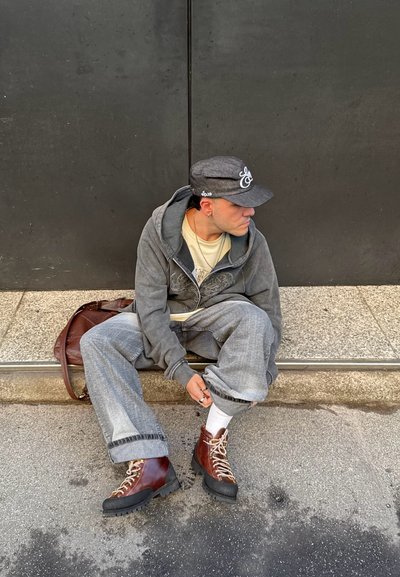 Persona con sudadera gris con capucha, vaqueros y botas marrones sentada en el bordillo, ajustando la pernera del pantalón, con gorra gris oscuro y calcetines blancos, con un bolso marrón al lado.