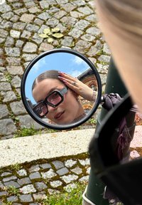 Czarne okrągłe lustro odbijające osobę w stylowych czarnych okularach przeciwsłonecznych, z gładkimi włosami i paznokciami pomalowanymi na jasnofioletowo. Tło z kostki brukowej.