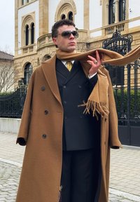 Hombre con abrigo marrón, bufanda a juego, traje negro y gafas de sol, de pie cerca de un edificio ornamentado y una reja de hierro forjado en la acera.