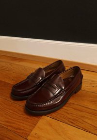 Bordo krāsas ādas loaferi ar dekoratīvu šuvi un gludu virsmu. Tiem ir zems papēdis un divi horizontāli siksnas akcenti.