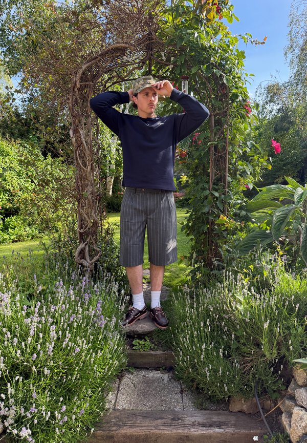 Joven ajustándose una gorra de camuflaje, de pie bajo un arco de jardín rodeado de plantas verdes y flores en un día soleado.