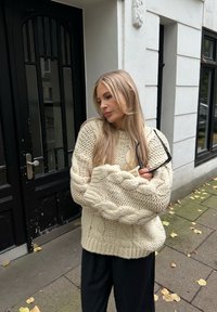 Suéter grueso de punto de cable en color crema con patrón texturizado y dobladillo acanalado, usado sobre pantalones negros. Persona sosteniendo gafas de sol, de pie frente a un edificio.