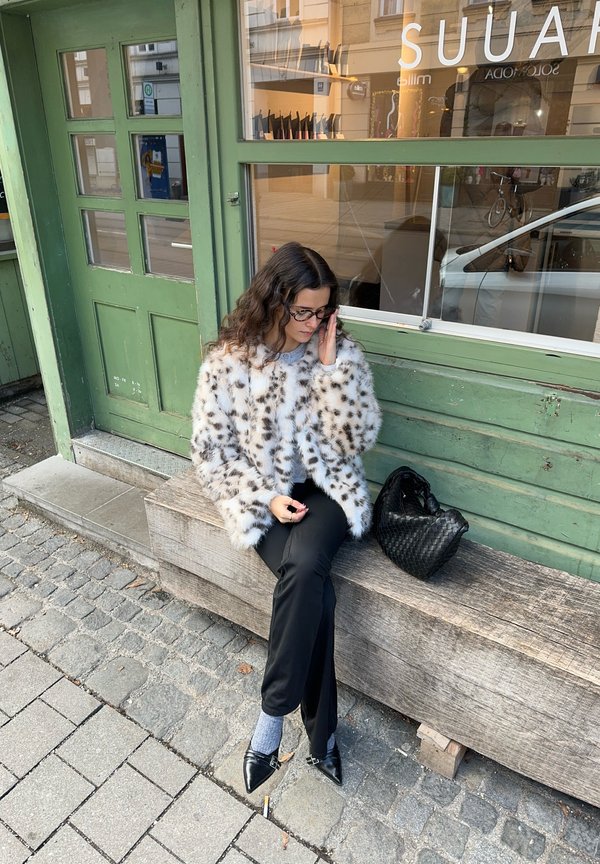 Faux fur jakke i hvid med brune pletter, kombineret med sorte bukser og grå sokker. Sort vævet taske og træbænk mod en grøn væg.