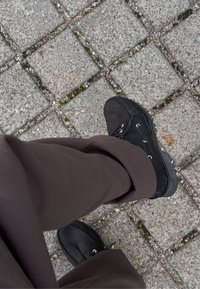 A person dressed in dark brown trousers and black lace-up shoes is standing on a grey stone pavement, which has moss growing in the gaps.