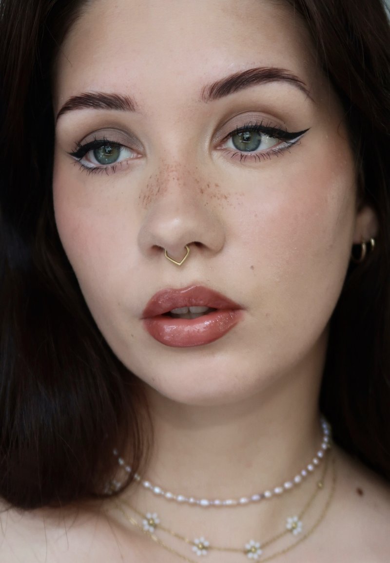 Jeune femme aux yeux verts, eye-liner ailé, taches de rousseur, un anneau de septum doré et des colliers superposés de perles et de fleurs.