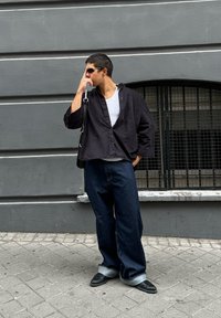 Un uomo con occhiali da sole, camicia nera, top bianco, borsa e pantaloni larghi scuri si trova sul marciapiede vicino a un muro grigio con una finestra con barre.