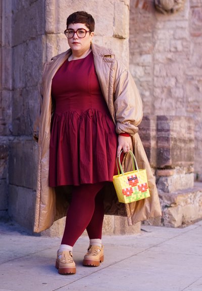Vestido burdeos con cintura peplum, abrigo acolchado beige de gran tamaño, bolso amarillo con estampado, medias rojas y zapatos de plataforma beige con detalles.