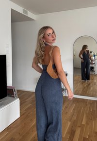 Marinblå ribbad jumpsuit med djup rygg, kombinerad med en tanfärgad mocka crossbody-väska med knuten rem. Trägolv och vita väggar i bakgrunden.