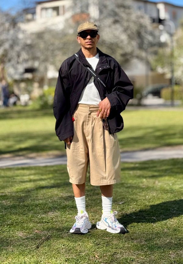 Homme portant une casquette beige, une veste noire, une chemise blanche, un short long beige, des chaussettes blanches et des baskets colorées, debout sur l’herbe dans un parc ensoleillé.