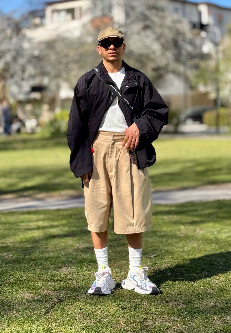 Homme portant une casquette beige, une veste noire, une chemise blanche, un short long beige, des chaussettes blanches et des baskets colorées, debout sur l’herbe dans un parc ensoleillé.