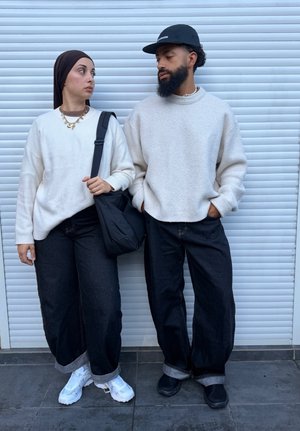 Dos personas con suéteres blancos y pantalones negros están de pie una al lado de la otra frente a una persiana metálica blanca, mirándose mutuamente.